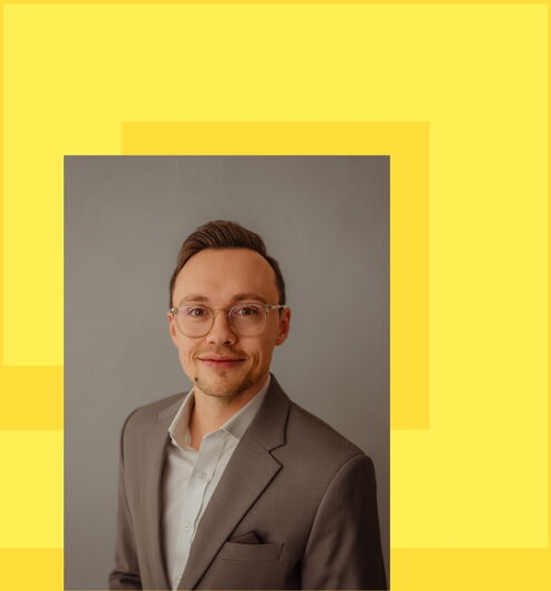 Porträt eines jungen Mannes mit Brille: Er sitzt entspannt auf einem hellgrauen Sofa, trägt ein weisses Hemd, dunkle Hosen und weisse Sneaker. Der Hintergrund ist neutral und hell, während ein gelbes, grafisches Designelement mit überlappenden Rechtecken